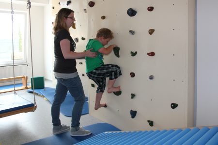Kind beim Klettern an einer Kletterwand in einem Raum mit einer Physiotherapeutin als Unterst&uuml;tzung