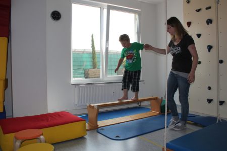 Kind balanciert mit Hilfe einer Physiotherapeutin auf einem Balken in einem Trainingsraum