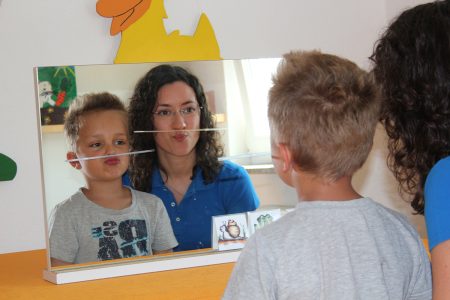 Ein Logop&auml;de und ein Kind schauen in den Spiegel, &uuml;ben Gesichtsausdr&uuml;cke