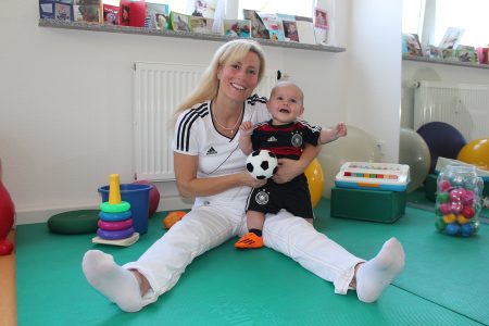 L&auml;chelnde Physiotherapeutin mit Baby und Stoff-Fu&szlig;ball