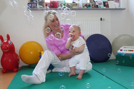 Physiotherapeutin spielt mit lachendem Baby auf der Matte mit Seifenblasen und B&auml;llen